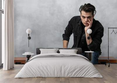 Studio fashion portrait of attractive young man in black jacket and blue jeans sitting against grey textured wall. Guy touches temples with fingers in thoughts Wall mural