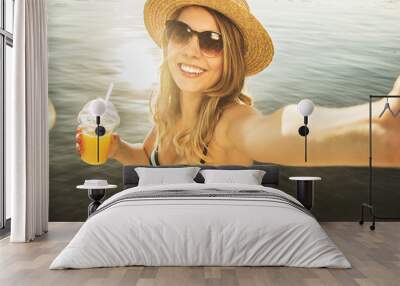 Capturing every happy summer moments. High-angle view of beautiful and young blonde haired woman keeping camera in hand and smiling while taking selfie with orange juice on the beach. Wall mural