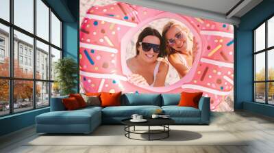 Cheerful beautiful young women friends look through inflatable doughnut ring and smile while sit on beach. Attractive stylish girls in sunglasses sunbathing and posing with donut. Summer vacation Wall mural