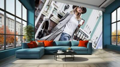 Coffee on the go. Stylish and young woman in coat and hat drinking coffee outdoors. Beautiful hipster girl in modern urban outfit walking city streets with paper cup of coffee. City fashion concept Wall mural