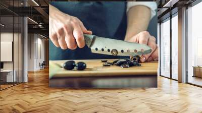 The chef in black apron cuts black olives with a knife. Concept of eco-friendly products for cooking Wall mural