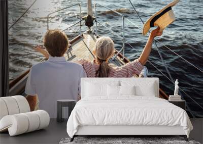 Back view of a happy mature woman waving her straw hat on a yacht. Senior couple enjoying a vacation on a sail boat. Wall mural