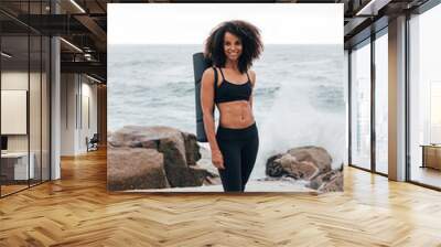 Beautiful smiling woman standing with yoga mat by ocean and looking at camera Wall mural