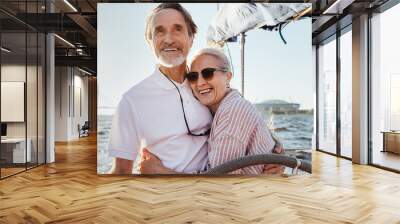 Cheerful senior couple standing at steering wheel on a yacht and hugging each other. Happy people enjoying sunny day on a boat. Wall mural