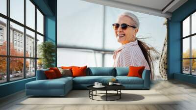 Laughing mature woman wearing sunglasses and looking at camera. Smiling female in casuals enjoying vacation. Wall mural