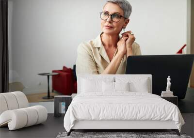 Thoughtful mature woman wearing eyeglasses sitting at a desk at home looking away Wall mural