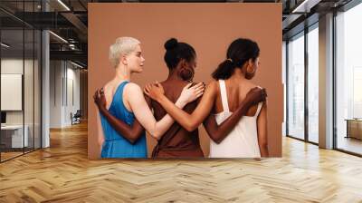 Three diverse women standing against brown background hugging each other Wall mural