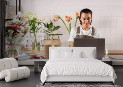 Young flower shop owner using laptop at counter. Woman florist standing at table.  Wall mural