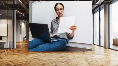 Young woman sitting on floor at kitchen counter with document in hand and using laptop Wall mural