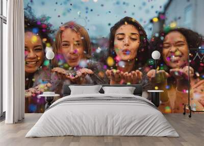 young women blowing confetti from hands. friends celebrating outdoors in evening at a terrace. Wall mural