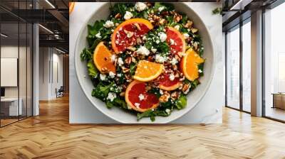 Top-down view of a bowl of bitter greens and citrus salad, with a sprinkle of feta cheese and nuts, set on a light marble surface. Wall mural