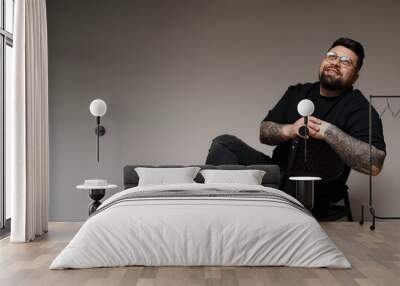 A photo of a tattooed man sitting on a black chair with his legs crossed. He is wearing a black t-shirt and black jeans. This image can be used for advertising, editorial, or personal projects. Wall mural