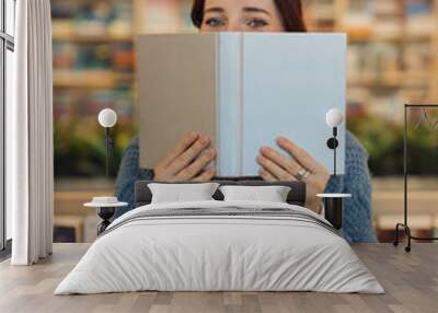 A young red-haired woman covers her face with a book in a cozy library setting, surrounded by shelves filled with books. Wall mural