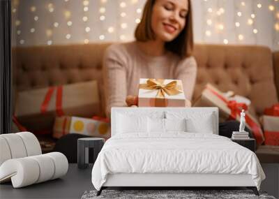 Happy young woman holding a present showing surrounded by gift boxes sitting crossed legs on a camel sofa with lights in background Wall mural