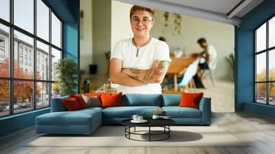 Confident transgender male with tattoos stands in a modern office, smiling colleagues working in the background. He embraces diversity, inclusion in the workplace, wearing casual attire and glasses. Wall mural