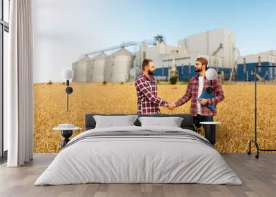 People shaking hands in a wheat field, farmer's agreement. Grain elevator terminal on background. Agriculture agronomist business contract concept Wall mural