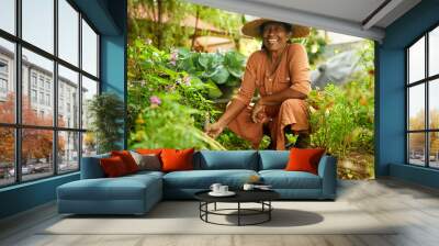 Senior happy Indian female farmer in straw hat sitting in garden taking care of plants. Elderly Sri Lankan smiling woman in her orchard planting flowers into soil. Gardening concept. Happy retirement. Wall mural