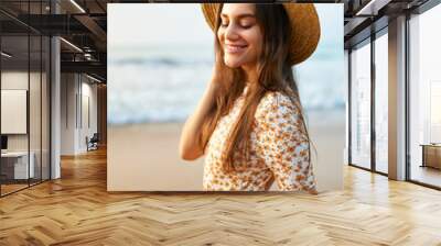 Smiling young woman in floral dress and straw hat stands on sandy beach at sunset. Relaxed female enjoys summer breeze by sea. Vintage style vacation mood. Happy tourist on coastal getaway. Wall mural