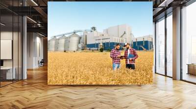 Two farmers stand in a wheat field with tablet. Agronomists discuss harvest and crops among ears of wheat with grain terminal elevator on background Wall mural