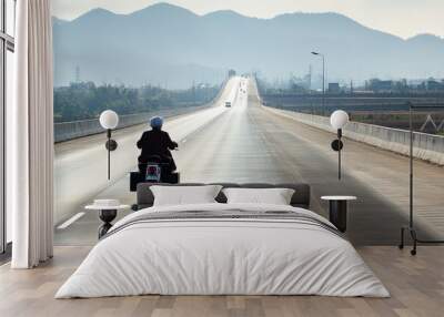 Daytime Photograph of Man Riding Motorcycle on Large Highway in Asia, Captured from Behind with Casual Attire and Helmet, Navigating Expansive Road Under Clear Daylight with Distant Hills and Greenery Wall mural