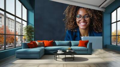 A businesswoman in a blue suit smiling while working on a laptop at the office. Portrait of a happy middle-aged woman with curly hair wearing glasses using a computer for work or online learning Wall mural