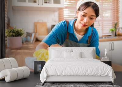 Professional Asian woman cleaning service wearing yellow rubber gloves, using a rag to wipe with spraying liquid detergent on the wooden top kitchen counter at home. Housekeeping cleanup, cleaner. Wall mural