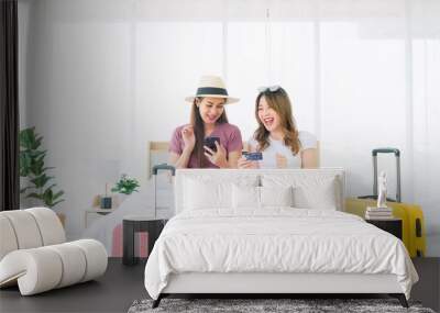 Two happy Asian women booked a hotel room on a smart phone and pay with a credit card while sitting together on the bed with their luggage. Planning on holiday vacation trip. Image with copy space. Wall mural