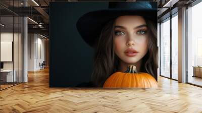 Young woman in black dress and witch hat holds pumpkin against dark background, close up. Mysterious witch and Halloween symbol, spooky celebration. Banner, party invitation, greeting card, copy space Wall mural