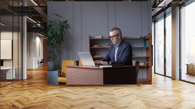 Mature pensive businessman with grey hair in glasses and suit is working on laptop in office Wall mural