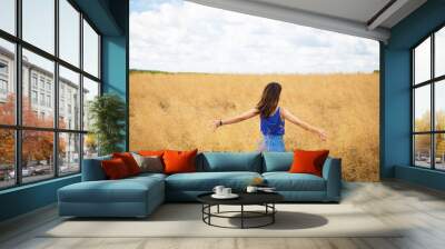 Portrait of a young beautiful brunette woman in a blue T-shirt and denim shorts stands in the middle of the field, exposing her face to the sun on the background of a summer day outdoors. Wall mural