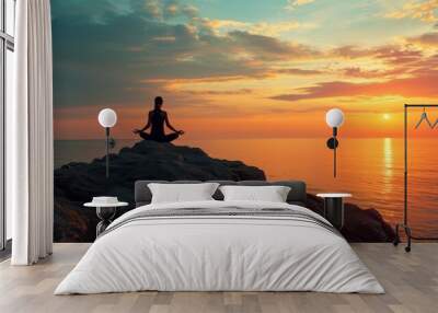 Silhouette of a person meditating in lotus pose on a cliff overlooking the sea at sunset. Wall mural