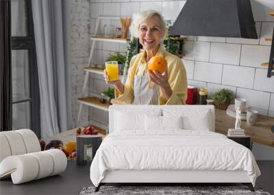 Attractive senior woman drinking freshly squeezed orange juice, looking happy and healthy. Concept of active lifestyle of mature people, cooking organic food. Mature female at home at her kitchen Wall mural