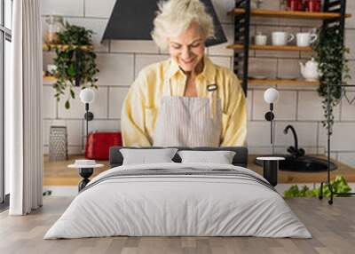 Attractive senior woman with grey hair cooking healthy food on her kitchen at home. Mature female preparing fresh vegetarian salad with organic ingredients from the market: tomato, cucumber, pepper Wall mural