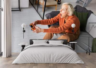 Attractive senior woman with grey hair playing console at home, having fun. Concept of mature female using technology, active lifestyle of pensioner, enjoying life Wall mural