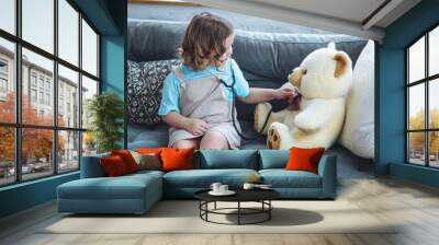 Cute little girl in a beautiful dress paying doctor at home with her teddy bear, listening to heartbeat with a stethoscope. Cozy atmosphere, kid's leisure and developing games Wall mural