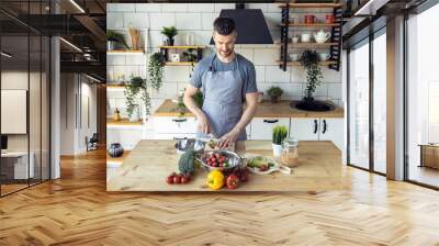 Handsome father, strong young man cooking healthy vegetable salad with fresh organic ingredients, tasty food in the kitchen at home . Men doing chores. Ripe pepper, tomato, cucumber Wall mural