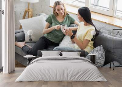 Two millennial girl friends at home on the sofa talking, drinking coffee, discussing. Diverse friends spending fun time together and laughing, chat and talk. Asian and Caucasian model. Wall mural