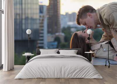 Young happy beautiful loving couple on a surprise romantic date on a rooftop on Saint Valentine's Day. Attractive man and woman, male and female hugging, kissing. Sunset, skyscrapers urban view banner Wall mural