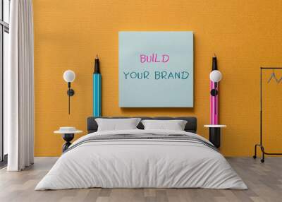 Text sign showing Build Your Brand. Business photo showcasing enhancing brand equity using advertising campaigns Front view blank colored sticky note two ballpoints cool yolk background Wall mural