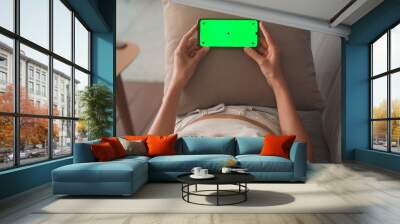 Woman lying down on couch and holding smartphone with green screen in horizontal landscape mode Wall mural