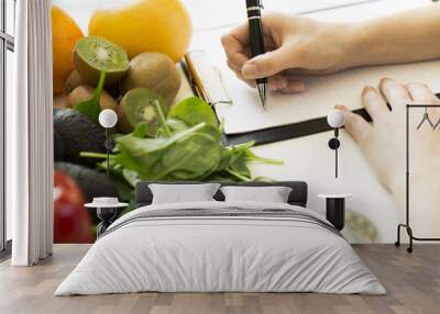 Nutritionist woman writing diet plan on table full of fruits and vegetables Wall mural