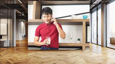 Smart, young and healthy Asian man holding a glass of milk. Young man using smartphone in the loft style kitchen room Wall mural