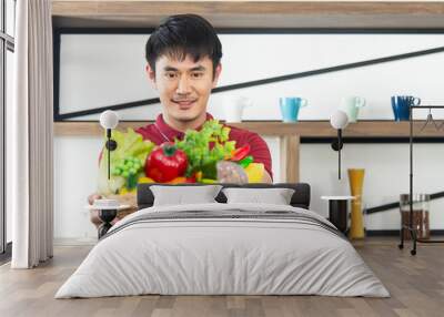 Smart, young and healthy Asian man with casual  red t-shirt holding  and show vegetable basket.  Young man cooking food  in the loft style kitchen room Wall mural