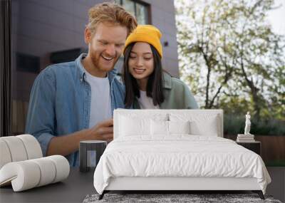 Couple of smiling stylish friends holding credit card, using mobile phone for online shopping. Asian woman and caucasian man ordering food  Wall mural