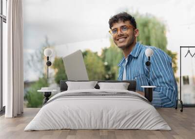 Portrait of handsome Indian copywriter using laptop computer, typing, working freelance project online, sitting outdoors. Smiling asian student studying, learning language, online education concept Wall mural