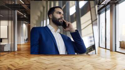 Portrait of handsome pensive middle eastern businessman talking on mobile phone looking at window standing in modern office Wall mural