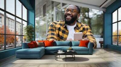 Young handsome African American man in stylish eyeglasses holding mobile phone, communication. Portrait of emotional guy using mobile application for online shopping, laughing, sitting in cafe Wall mural