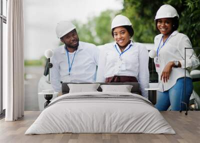 African american technician in white helmets near car. Group of three black engineers meeting. Wall mural