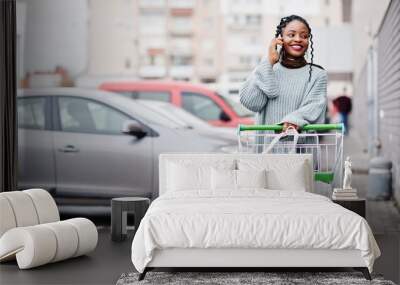 African woman with shopping cart trolley posed outdoor market near car parking and speaking on phone. Wall mural