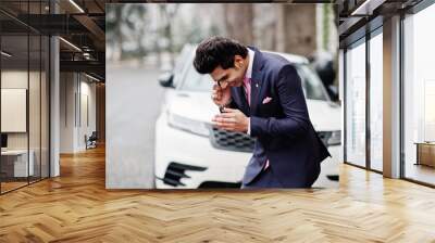 Elegant indian macho man model on suit and pink tie posed against white business car and speaking on phone. Wall mural